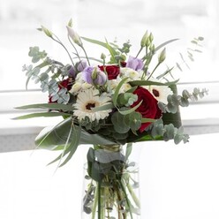 In the softly bright ambiance that calls to mind quiet mornings in Gerrards Cross, this bouquet balances confidence and poise. The deep, plush reds of each rose command the eye, softened by the radiant white of plump gerberas, complete with cocoa-brown centers that lend a vintage charm. Throughout, sprigs of round-leaf eucalyptus form a meandering, silvery-green frame, their subtle aroma gently reminiscent of the nearby Bulstrode Park woods. The lisianthus, in shades of gentle lavender, rest lightly amidst the main blooms, some buds just beginning to open-each one a symbol of promise, much like the hope that blooms with every new season along Packhorse Road. Voluminous, cloud-like white filler flowers add layers of softness, and the bouquet is enveloped by broad, emerald foliage that shines against the clear, pure background. The glass vase, understated yet elegant, reveals the tangle of strong green stems resting in cool water, underscoring the arrangement's freshness. Whether chosen to celebrate intimate family gatherings or to brighten homes from Chalfont St Peter to Gerrards Cross, this bouquet expresses comfort, elegance, and the thoughtful craftsmanship of a local florist deeply attuned to community and occasion.