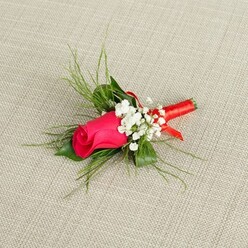 A romantic boutonnière is displayed on a pale, linen-like fabric background, its subtle beige tones gently textured like the table linens often used for weddings in Gerrards Cross hotels and countryside barns. The focal point is a single, luscious rosebud in a vivid fuchsia or deep pink shade, still tightly furled so that each petal curves inward, catching the light with a soft satin sheen. Nestled close against the rose are clusters of tiny white gypsophila, or baby's breath, their airy, frothy blossoms creating a delicate halo that both softens and highlights the bold hue of the central flower. Surrounding this are slender sprigs of light, feathery green foliage, fanning outward in fine threads that bring a sense of movement and garden freshness, as if just gathered from a well-kept flower bed along one of the leafy streets leading down from the station. The stems at the base of the arrangement are tightly bound with a narrow, coral-to-red satin ribbon, wound with precise, florist's care so that no ends stray, giving the impression of a handcrafted piece made to be worn with pride. The lighting is bright and clear, revealing every texture: the smooth rose petals, the tiny star-like gypsophila blooms, the almost feathery greenery, and the gentle weave of the fabric beneath. This single-stem buttonhole feels like the finishing touch for a wedding morning, a prom at a local school, or a special event where the wearer wants a subtle but heartfelt nod to tradition, love, and thoughtful floral design.