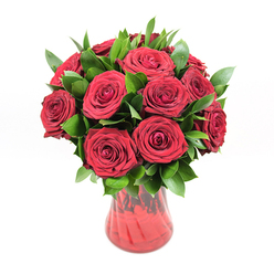 A classic dozen-plus deep red roses rise in a graceful cluster from a translucent red glass vase, set against a pure white backdrop that makes every shade and texture clearly visible. The roses are at that perfect stage of bloom a professional florist looks for: fully opened to reveal their lush, velvety spirals, yet still firm and confident, promising several days of beauty. Each petal curves outward in smooth arcs, creating a rich tapestry of crimson that feels both luxurious and intimate. Dark green foliage, likely ruscus, is carefully threaded between the blooms, the pointed, almond-shaped leaves creating a rhythm of glossy green that frames the roses without overwhelming them. The foliage adds depth and shadow, making some roses appear to sit forward while others recede, giving the arrangement a three-dimensional, sculpted presence. The vase itself is a deep red glass with a gently flared opening, a softly cinched waist, and a rounded base, its subtle ribbed texture catching the light so that darker and lighter streaks of ruby seem to ripple down its sides. This interplay of glass and bloom creates a harmonious monochrome palette, united by the green accents. Because the background is bright white and evenly lit, the bouquet appears almost to glow, like a centerpiece ready to be placed on a dining table in a Gerrards Cross townhouse off Packhorse Road. The overall feeling is one of sincere, undiluted emotion: this is the sort of rose arrangement chosen for anniversaries, romantic birthdays, or a surprise delivery across Gerrards Cross when someone wants their love to be understood without a single word.