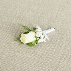 A refined wedding boutonniere is displayed on a pale beige, linen-like fabric that runs diagonally beneath it, creating a calm, understated stage for the floral detail. At its centre, a single creamy white rosebud is captured in that fleeting moment just before it opens fully, its petals smooth, velvety, and gently cupped, like a promise about to unfold. The rose is surrounded by airy sprays of white baby's breath, each tiny floret glowing softly in the diffused light, giving the arrangement an almost cloud-like aura. Slender, deep green leaves and a few lighter, feathery fronds of foliage cradle the bloom, their shapes layered in a way that adds depth and texture without disturbing the boutonniere's compact silhouette. The stems are gathered together and wrapped tightly with a crisp white satin ribbon, which is finished with a neat, dainty bow sitting just beneath the main flower, suggesting the careful craftsmanship of a florist preparing for a ceremony in Gerrards Cross, perhaps just off Oxford Road or near the station. The gentle lighting highlights the contrast between the pure white petals, the intricate clusters of gypsophila, and the rich greens, while the discreet weave of the background fabric adds subtle interest without distracting from the flowers. The overall impression is of quiet elegance and thoughtful detail, the sort of boutonniere a groom might wear for photographs in West Common or along the leafy streets leading toward the Chilterns. It feels both personal and traditional, capturing the essence of a local celebration, where every small element, even a single rose on a lapel, is chosen to reflect love, care, and a sense of place.