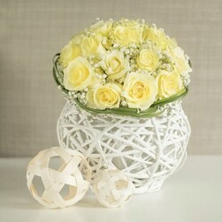 On a pristine white tabletop that could easily belong to a boutique venue near Gerrards Cross station, a romantic floral centerpiece becomes the calm heart of the scene. At its core, an abundant mound of pale yellow roses rises in a flawless dome, every bloom carefully positioned so their velvety petals overlap, creating a continuous field of buttery colour. The roses are at their peak, neither too tight nor too open, giving a sense of poised freshness as if just arranged by a local florist moments ago. Threading gently through the roses are wispy sprays of white gypsophila; the countless tiny flowers form a light, frothy halo, reminiscent of fine mist drifting across the Misbourne Valley. Around the lower edge of the bouquet, a slim band of polished green foliage has been wrapped with intention, adding a crisp ring of colour that marks the transition from petals to vase. Supporting this floral sculpture is an eye-catching white woven container, shaped in a rounded, slightly waisted silhouette. The open, basket-like structure of the vase allows the neutral background to show through its crisscrossing strands, playing with light and giving the whole arrangement a sense of airy modern design. Beside the vase, two spherical accents made from thin, natural rattan-like strips extend the woven theme, their pale tones and cage-like patterns subtly mirroring the container's texture. The softly blurred beige backdrop offers no distraction, simply a calm canvas that lets the gentle yellows, fresh greens, and warm whites radiate. The whole composition feels ideal for a wedding breakfast table in Gerrards Cross, a refined anniversary dinner, or a quiet celebration of love and new beginnings.