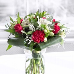 A lush, romantic bouquet is showcased in a tall, clear glass cylinder vase, the stems submerged in water and neatly trimmed so each line looks deliberate and refined. The main impression is a confident play of red and white, like a classic love letter translated into flowers. Deep red roses with velvety petals spiral open at the center, their edges soft and gently curling, while a richly textured crimson carnation sits at the front, its densely ruffled petals creating a focal point of movement and depth. Interwoven with the red blooms are creamy white flowers-elegant alstroemeria whose narrow petals show fine, painterly speckles, plus pure white blossoms and several closed white lily buds that stand tall and smooth, suggesting the scent that will emerge over the coming days. Light clusters of gypsophila float among the larger heads, their tiny white flowers like a dusting of snow, softening the colour contrasts and giving the design an almost ethereal haze. Surrounding everything, a generous spread of greenery forms a rich frame: broad glossy leaves are curled and wrapped tightly around the base, while layered eucalyptus-style foliage and pointed leaves offer cooler tones and added structure. The bouquet sits on a pristine white surface, backed by a softly blurred, light-filled background that suggests a bright room in Gerrards Cross, perhaps a dining space near East Common catching the afternoon sun. The atmosphere is polished yet warm, ideal for marking milestones, sending congratulations, or expressing deep affection with a same-day delivery from a trusted local florist to homes and offices across the area.
