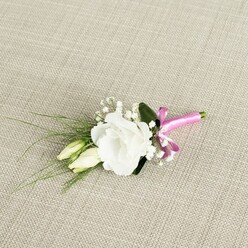 A finely detailed boutonnière rests at a gentle diagonal on a warm, oatmeal-toned fabric, whose visible weave recalls natural linen often seen in Gerrards Cross homes and boutique venues. The focal point is a pristine white lisianthus flower, fully open and luxuriant, its ruffled petals layered in soft concentric circles. The bloom feels almost weightless, the edges of each petal catching the light in a way that suggests both elegance and tenderness, as if it has just been picked fresh from a garden near Austenwood Lane. Just below and to the left of the main flower, two slender lisianthus buds lean close, their cream and pale green tones giving a sense of quiet anticipation, like the moments before a wedding ceremony begins. Fine sprigs of gypsophila weave through the design, their tiny white blossoms forming a delicate, cloud-like halo that softens the boundary between flower and foliage. From behind the bloom, narrow, grass-like green leaves fan out toward the left, adding texture and a slight sense of movement, while a single broader, darker leaf frames the lisianthus and lends a subtle depth of colour. The lower stems are gathered neatly and wrapped with a radiant fuchsia satin ribbon, tied into a small, tidy bow that injects a chic, contemporary pop of pink into the otherwise classic white-and-green scheme. The soft, even lighting allows every petal, bud, and leaf to be clearly seen, giving the whole piece an air of calm refinement. It feels like the perfect finishing touch for a groom or groomsman dressing for a ceremony in one of Gerrards Cross's churches or country house venues, a small floral accent that carries both style and sentiment.