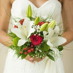 Imagine the gentle hush of a Gerrards Cross morning as a bride, radiant in a white, softly patterned gown, clasps a stunning bouquet crafted with artistry and heart. Crisp oriental lilies stand boldly at the centre, their pure white petals opening with grace, golden stamens glowing in the filtered light. Crimson red roses, each one a classic spiral of love, weave through the arrangement, their velvet richness grounding the bouquet in heartfelt romance. Vibrant red berries dot the gathering like festive notes, while unopened green lily buds hint at new beginnings and the unfolding journey ahead. Wisps of baby's breath drift between the larger blooms, lending the bouquet a sense of weightlessness-like a secret shared in the quiet corners of West Common or the gardens of East Common nearby. Mossy green button flowers and fine, fresh ferns provide an earthy base, their textures reminiscent of the local woodlands in spring. The composition feels alive, every petal and leaf thoughtfully placed to echo both tradition and joyful celebration. Held close in the bride's hands, the bouquet embodies hope, devotion, and the fresh promise of weddings in Gerrards Cross-a keepsake of beauty, memory, and community spirit.