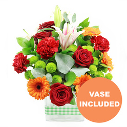 An exuberant, garden-party style bouquet is captured against a crisp white backdrop, overflowing with colour and texture in a way that feels perfectly suited to a joyful gathering in Gerrards Cross. At its core, several deep red roses stand proud, their velvety petals still partly furled, suggesting both romance and formality. These are echoed by plump, red-orange carnations with finely frilled edges, whose softer shapes bring warmth and a hint of playfulness, like laughter carried across West Common. Bright orange gerbera daisies radiate outwards, each bloom bearing a strong dark centre that anchors the vivid petals and draws the eye. Nestled between them, clusters of tiny, spherical lime green pompom chrysanthemums form little islands of colour, adding an upbeat, contemporary note that feels fresh and modern. Rising above this colourful crowd are stately white Oriental lilies, some dramatically open to reveal blush-pink veining and prominent stamens dusted with pollen, others still in sculptural bud, promising fragrant blooms in the days ahead. The flowers are supported by layered greenery: broad, glossy leaves create a dense base, while arching sprigs of silver-green eucalyptus introduce cool tone and a soft, airy outline. All the stems are gathered neatly into a modern, rectangular clear glass vase, which allows the clean lines of the stems to be seen and emphasises the bouquet's structured design. Around the lower half of the vase, a fresh light green and white gingham ribbon is wrapped in a tidy band, adding a homely, country-inspired detail reminiscent of local cafés and homes around Packhorse Road. In the bottom right corner of the image, an orange circular overlay with bold white lettering states "VASE INCLUDED," clearly showing that the stylish vase forms part of this ready-to-enjoy floral gift.