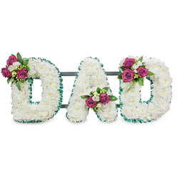 This carefully fashioned memorial reads clearly as the word "DAD", each letter sculpted from a dense carpet of immaculate white chrysanthemums whose fluffy florets create a soft, tactile surface that invites quiet reflection. An emerald green, ruffled ribbon trims both the outer and inner edges of each letter, its cool colour creating a crisp silhouette against the white blooms and lending a sense of composed dignity appropriate for a funeral setting. A discreet dark green or grey horizontal support runs behind the letters, suggesting the arrangement is ready to stand on an easel or be placed at a graveside service. Small clusters of accent flowers punctuate the composition: the first "D" wears its magenta rose cluster on the upper left curve, the "A" cradles its cluster low in the central V, and the final "D" balances the design with a matching cluster at the upper right. Those magenta roses - from bud to half-open bloom - are paired with delicate white freesia and rich green foliage, introducing warmth and depth without overwhelming the serene white field. Under gentle lighting the texture of the chrysanthemums and the satin sheen of the ribbon read clearly, while the mixed scents of rose and freesia add a tender note to the visual. As a local florist who prepares tributes for services near Gerrards Cross railway station, Bulstrode Park, and the High Street, Flower Delivery Gerrards Cross (flowerdeliverygerrardscross.co.uk) often offers this bespoke lettering as a heartfelt way to honour a beloved father at funerals and memorial gatherings across the area.
