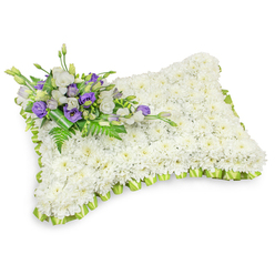 A serene rectangular floral pillow, crafted with the kind of attention we use daily at Flower Delivery Gerrards Cross, occupies a simple white setting where soft shadows give it depth and presence. The surface is a dense field of immaculate white chrysanthemum heads - hundreds of small blooms packed closely to create a uniform, cushion-like texture that suggests purity, rest and quiet reverence. The pillow's perimeter is finished with a pale lime-green ribbon, pleated into tidy overlapping loops that form a subtle scalloped border and introduce a gentle hint of optimism amid the solemn tones. Diagonally placed at the top-left, a composed spray of accent flowers provides emotional punctuation: white lisianthus with their soft, ruffled cups sit alongside rich purple lisianthus, some still in tight bud, their tones suggesting dignity and remembrance. Sprays of feathery fern and glossy green foliage thread through the cluster, offering contrast in both colour and texture and filling the design with a fresh vegetal perfume. The base of the accent grouping is wrapped with darker green leaves to secure the stems and give the piece a polished finish. Presented against a bright white background with a subtle drop shadow, the tribute reads as both an intimate keepsake and a dignified public offering suitable for local church services, graveside visits or memorials across Gerrards Cross, Chalfont St Peter and Beaconsfield. It speaks with quiet confidence - a tactile, visual message of sympathy when words fall short.