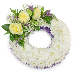 A dignified, hand-arranged circular funeral wreath set against a pure white studio background, crafted to convey calm and lasting remembrance. The main body is a dense, continuous ring of small white chrysanthemums, each head layered with ruffled petals to form a soft, cushiony surface that reads as serene and consoling. Nestled along the upper-left arc is a considered spray of accent flowers: three creamy yellow roses open into gentle rosettes, surrounded by clouds of white baby's breath (gypsophila) and sparing sprigs of lavender-purple statice or limonium that introduce a quiet accent tone. Beneath and around these blooms, varied green foliage - from broad, waxy leaves to delicate, fern-like fronds - provides sculptural support and a fresh, verdant backdrop. A subtle purple mesh or ribbon is visible beneath the chrysanthemums at both the inner and outer rims, a refined detail that adds depth and a discreet note of colour. The wreath's open centre remains uncluttered, symbolising continuity and the circle of life; soft, diffused lighting highlights the textural contrast between the velvety roses and the feathery chrysanthemum field while casting a gentle shadow that gives the piece presence on the white plane. The composition reads as comforting and unembellished - suitable for services across Gerrards Cross, Seer Green and nearby villages - and has been prepared with the care and craftsmanship expected from Flower Delivery Gerrards Cross for families arranging memorials and tributes.