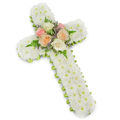 A beautifully crafted floral cross for a funeral or memorial is displayed against a stark white studio background, the soft shadow beneath it giving the impression of a three-dimensional piece resting lightly in space. The entire cross is packed with flawless white button chrysanthemums, their tiny petals forming a dense, cushion-like texture that covers every inch of the design. This carpet of snowy blooms feels serene and contemplative, echoing the quiet stillness of a reflective walk through Gerrards Cross Common. At the heart of the cross, where the vertical and horizontal arms intersect, a small but striking bouquet of roses becomes the emotional centre. Open-faced roses in gentle tones of blush pink, ivory, and warm peach are cradled together, each bloom unfurling in tender layers. Among them, feathery sprigs of lavender-hued filler flowers and fine green fern fronds add subtle movement and contrast, softening the transition between the crisp white base and the pastel blossoms. Encircling the whole shape, a slim lime green ribbon traces the outline of the cross, just visible beneath the chrysanthemums like a fresh spring border, offering a whisper of colour and renewal. The overall impression is one of calm reverence and heartfelt love, a thoughtful tribute that could be placed beside a coffin, near a chapel altar, or at a graveside in the Gerrards Cross area. Every flower appears carefully chosen and positioned, reflecting the steady hands and compassionate heart of an experienced local florist who understands how flowers can offer comfort when words are hard to find.