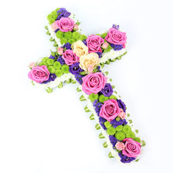 Set against a spotless white background, this cross-shaped funeral tribute is captured from a slight overhead angle, allowing its thoughtful floristry to unfold in layers. A neat border of tiny white spray chrysanthemums runs around the full outline of the cross, their close-packed petals forming a soft, cushion-like frame that suggests purity and peace. Just beneath, a slim band of folded light green ribbon or foliage encircles the shape, adding a refined edge and a hint of fresh spring colour. Inside this border, the cross is richly filled with a tapestry of blossoms: bold, fully opened roses in hot fuchsia pink and gentle creamy yellow create luminous focal points along the arms and centre. These roses are interwoven with bright lime-green button chrysanthemums, whose small, rounded heads bring playful texture and a contemporary feel to the classic design. Nestled between them, clusters of deep purple and violet blooms, probably lisianthus or statice, add depth and a slightly perfumed, contemplative mood. The cross is tilted subtly from top left to bottom right, giving it a dynamic, almost floating presence, as though it might be resting in quiet remembrance at a service in a Gerrards Cross church. Clean, bright lighting ensures each colour and petal is clear and true, from the crisp whites to the rich purples. The arrangement feels both respectful and uplifting, a floral cross that speaks of faith, enduring love, and gentle comfort, suitable for same-day sympathy delivery to local chapels, crematoriums, and homes throughout Gerrards Cross.