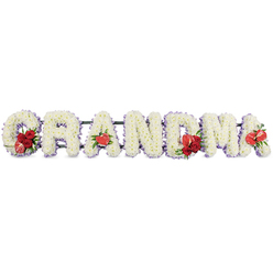 The image shows a beautifully composed funeral flower tribute that spells out GRANDMA in large, block capital letters, arranged in a straight line as though ready to be placed beside a coffin or near the lectern at a memorial in Gerrards Cross. Every letter is completely blanketed in tightly packed white chrysanthemums, their small star-shaped florets pressed closely together to create a plush, cushion-like effect that looks both soft and pristine. The white blooms are framed by a subtle edging of delicate purple or lilac ribbon, which peeps out around each letter like a soft outline, lending a hint of colour and a restful, almost spiritual glow. Scattered with intention along the length of the word are vibrant red floral groupings that break the white with moments of warmth and passion. At the opening G, a generous cluster of deep red roses is paired with an unusual variegated red-and-white flower, likely a calla lily, setting a tone of love and individuality right from the start. The first A features a single, polished red anthurium lily, heart-shaped and slightly waxy, resting at the base like a symbol of devotion. In the middle, the M is adorned with more red roses and another red anthurium, echoing the same heartfelt note, while the last A is crowned with a final red anthurium, bringing a gentle sense of balance and completion. All the letters sit on a discreet dark green metal or foam frame that holds them upright while almost disappearing from view. The overall tribute feels serene, respectful, and deeply affectionate, the kind of bespoke GRANDMA arrangement a local Gerrards Cross family might choose for a funeral at a nearby crematorium or a quiet graveside farewell.