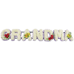 The image shows a lovingly crafted GRANDMA funeral flower tribute laid out in a long horizontal line, the letters standing bold and clear against a bright, uncluttered white background. Each capital letter is formed from a tightly packed bed of small white chrysanthemums, so dense they create a plush, cushion-like surface with a soft, almost luminous glow. Beneath the white blooms, a neat edging of pale lavender flowers peeks out, tracing the lower edge of every letter and adding a gentle veil of colour that gives the word depth and definition without disturbing its peaceful simplicity. A few carefully placed accent clusters bring the tribute to life. On the lower left of the 'G', three velvety, deep red roses are tucked into rich green foliage, a striking symbol of devotion and enduring love. The first 'A' carries two yellow-green cymbidium orchids with speckled, reddish-brown throats, resting elegantly across its crossbar and lending an air of quiet luxury. The central letters 'R', 'N', and 'D' remain purely white and lavender-edged, representing peace, innocence, and calm reflection. Towards the right, the 'M' is graced with another trio of red roses in its lower right dip, echoing the first accent of the design, while the final 'A' repeats the twin orchids, bookending the tribute in symmetry. A slim, dark green metal frame discreetly links the letters along the top, ready to rest on trestles in a church, crematorium, or graveside in and around Gerrards Cross. The overall mood is dignified and heartfelt, like a final handwritten note translated into flowers, offering comfort and a focal point for memories of a cherished grandmother.