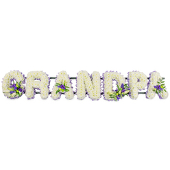 A heartfelt memorial arrangement spells out the word GRANDPA in large, upright block letters, laid out horizontally as though placed along the side of a coffin or at the front of a service in Gerrards Cross. Every letter is completely covered with clusters of small white chrysanthemums or carnations, each bloom carefully set close to the next so that no gaps show, forming a smooth, plush surface that feels both tender and dignified. The white petals catch the light softly, suggesting purity, gratitude, and quiet remembrance. Among this sea of white, the florist has woven in thoughtful touches of purple and lavender flowers, perhaps lisianthus or dainty chrysanthemums, which appear in small, intentional groupings: at the base of the opening G, inside the hollow of the first A, nestled in the second A, and accenting the curves of the P. These richer tones bring depth and a soothing, almost spiritual note to the design, while hints of dark and mid-green foliage add fresh, leafy texture. A discrete dark green support bar runs along the bottom, mostly hidden under the blooms, subtly linking each floral letter into a single, unified tribute. The overall impression is of a lovingly crafted funeral piece that a family from Gerrards Cross might arrange for a final farewell, perhaps after gathering in nearby East Common or returning from a service at a local church. Its gentle balance of colour, lush texture, and careful craftsmanship speaks of the bond between generations and the enduring impact of a beloved grandfather's life.