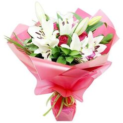 A striking bouquet of fresh white Oriental lilies and deep red roses is shown in close detail, styled to look as if it has just been prepared on the workbench of a skilled Gerrards Cross florist. The lilies dominate the composition, their large, trumpet-shaped flowers arching outward with a sense of poise, each petal a soft, luminous white that gently curls at the tips. Inside the blooms, dark reddish-brown anthers sit on slender filaments, dusted with pollen that contrasts dramatically with the pure petals. Tucked among the open flowers are smooth, tapered lily buds in shades of green and cream, hinting at future blossoms and long-lasting enjoyment once the bouquet is delivered to homes around West Common or the leafy streets near Fulmer Road. Between the lilies, tightly furled red rosebuds and slightly open roses provide deep, velvety pools of colour, their romantic tone balanced by layers of rich greenery. Broad, shiny leaves and fine, spiky blades stretch and curve around the blooms, forming a lush, full framework that suggests abundance rather than clutter. The stems are gathered into a generous wrap of bright pink, semi-translucent cellophane, which forms soft waves and folds that catch the light like ripples on a festive ribbon. Around the base, a slim lime green satin bow secures the wrapping, its fresh, zesty hue echoing the foliage and adding a playful accent. The overall impression is of a joyful, sophisticated gift perfect for celebrating anniversaries, promotions, or special family gatherings in Gerrards Cross, radiating affection, gratitude, and a quiet sense of occasion.