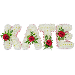 This handcrafted memorial display shapes the letters K, A, T and E from an enveloping mass of tiny white chrysanthemums, each bloom pressed closely so the letters read as soft, rounded forms with a cloud-like volume on a plain white backdrop. A pale pink satin ribbon traces each letter's edge, a refined border that warms the composition and gives the silhouette a finished, thoughtful line. Each letter is punctuated by a single, velvety deep red rose framed by abundant green foliage - a mix of broad, glossy leaves and feathery fern sprigs - which adds texture, depth and movement to the otherwise serene white field. The placement of each rose is deliberate: on the K it is set along the lower part of the left vertical stroke, the A's rose nestles at the top peak, the T's bloom is positioned at the base of the left side of its horizontal crossbar, and the E is highlighted at its middle horizontal bar. The red roses lend a warm, peppered fragrance that softly contrasts with the clean chrysanthemum scent; the satin ribbon catches a quiet shine under soft lighting, and the foliage brings a living resilience to the piece. As a local florist working in Gerrards Cross and the surrounding villages, I design name tributes like this to convey love, purity and remembrance at funerals or commemorative services, balancing clear lettering and tactile detail so the tribute reads well from the front pew and invites closer reflection for those who walk beside it.
