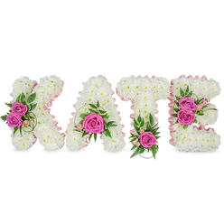 Handcrafted with quiet care, this personalised floral name tribute spells KATE in large, freestanding letters built from hundreds of small, immaculate white chrysanthemums that form a dense, cloud-like texture. A delicate pale-pink satin ribbon outlines the inner contours of each letter, subtly framing the shape and giving a soft blush against the pristine white. Hot pink roses punctuate the design - two on the K, a central bloom within the A's lower loop, one anchoring the T, and a clustered pair on the E's right side - their vivid colour and soft perfume drawing the eye and lending a compassionate warmth. Interspersed are delicate pale pink buds and mixed greenery, including ferny fronds and glossy foliage, which tuck around the roses and lift the composition with natural movement. The letters cast slight shadows on the white surface beneath, creating a gentle three-dimensional presence suited to a funeral easel or a celebration-of-life display. The overall effect is quiet, dignified and intimately personal: white for purity and remembrance, pink for affection and gentle celebration. As a Gerrards Cross florist I often prepare arrangements like this for services at local chapels, private memorials on the High Street or floral tributes placed near the station; Flower Delivery Gerrards Cross can adapt the size and rose placement to reflect the personality of the person being honoured.