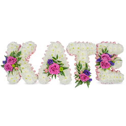 A lovingly created memorial display reads 'KATE' in four upright letters, each letter painstakingly formed from a dense layer of immaculate white chrysanthemums that create a smooth, cushioned surface. A soft pastel pink satin ribbon outlines the shapes, adding an understated warmth and a subtle sheen under diffused light. Each letter is punctuated with carefully placed accent clusters: large, velvety fuchsia-pink roses, sprays of delicate pale pink roses, and small deep purple or blue blooms - possibly statice or lisianthus - which together bring depth and a comforting colour contrast to the white base. Floral placements are intentional and balanced: the diagonal stroke of the 'K' hosts a prominent spray; the 'A' is brightened near its lower right curve; the 'T' is grounded with a bloom cluster at its foot; and the 'E' carries two groupings along its middle and bottom bars. Interspersed among the flowers are eucalyptus leaves, soft fern fronds and mixed greenery that add texture, a faint herbal scent and a sense of natural movement. As an experienced local florist, Flower Delivery Gerrards Cross would present this piece with sensitivity for a funeral, wake or celebration of life at venues such as the church near the station or a chapel at one of the nearby crematoria. The design balances purity and vivid memory: the white chrysanthemums signify remembrance while the pinks and purples express love and gratitude, making this a calming, personalised tribute for family and friends.