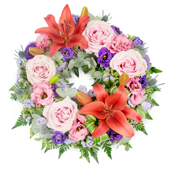 A beautifully balanced sympathy wreath is photographed from above against a clean white background, its circular form symbolising eternity and unending affection. Two prominent coral-orange Asiatic lilies, one at the upper curve and another towards the lower edge, create strong focal points; their broad petals curl gracefully, revealing dusted stamens that draw the eye and add a sense of warmth amid the softer tones. Surrounding them, generous clusters of blush and lavender-pink roses are arranged in a natural rhythm, some fully open and others just beginning to unfurl, their velvety spirals conveying gentleness and quiet grace. Between the roses, ruffled lisianthus blossoms in pale pink and rich purple weave through the design, their cup-shaped flowers adding a romantic, almost cloud-like texture. Scattered throughout are delicate sprays of tiny lavender-blue filler blooms, likely statice, that give the impression of a soft haze resting over the wreath. The base is lush with textured greenery: rounded, silvery eucalyptus leaves, slender fern fronds, and spiky blue-grey eryngium thistles that introduce a slightly rustic, countryside note reminiscent of the hedgerows around Gerrards Cross and its leafy lanes. Several closed lily buds nestle quietly in the foliage, promising future colour and symbolising memories still cherished. A few bright green, folded foliage accents at the base of the lilies add a contemporary flourish, hinting at the skilled hands that shaped this design. Created by local florists who understand the tone of services held in and around Gerrards Cross, this funeral wreath feels calm, respectful, and deeply personal, suitable for honouring a loved one at a chapel, crematorium, or graveside gathering.