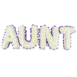 A lovingly composed memorial arrangement spells AUNT in bold, elegant capitals, each letter formed from hundreds of small, pristine white spray chrysanthemum flowers whose delicate petals create a cushiony, cloud-like surface. Here and there the pale yellow centres peek through, softening the white with a natural, sunlit warmth. The perimeter of every letter is neatly outlined with a rich purple satin ribbon, the fabric pleated into a scalloped edge so the ribbon's glossy face plays against the matte petals. Low grey shadows beneath the letters emphasise their raised structure against the clean white field, lending the piece a calm, sculptural presence. There is a faint, fresh scent-green and dewy-typical of newly gathered chrysanthemums, and the arrangement conveys a tone of respectful celebration rather than stark mourning. As a local florist familiar with Gerrards Cross, I picture this tribute placed near the war memorial on the High Street or within a family service at a chapel close to the station; it is the kind of personalised tribute we craft for neighbours and friends. Flower Delivery Gerrards Cross would prepare this with the same care shown here: secure framing, long-lasting blooms, and ribbonwork that provides dignity and a gentle pop of colour to honour a cherished aunt.