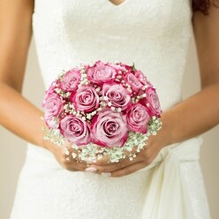 Soft, natural daylight illuminates this intimate bridal moment, as seen through a close-up of a bride's hands gently embracing a perfectly round bouquet set against her lace-clad white gown in Gerrards Cross. Each rose radiates with almost painterly precision, their centers opening in gentle blush tones before deepening to vibrant fuchsia at the outermost petals-a seamless transition reminiscent of the sunsets over East Common Park. Babbling alongside the blooms are frothy tufts of baby's breath, adding a weightless, ethereal quality that lightens the composition and recalls the fresh, pure air of the local countryside. The hands, warm-toned and beautifully manicured in pale pink, cradle the bouquet with measured care. Above them, the bodice of the dress reveals a pattern of raised, floral lace, edged by an elegant V-neckline and complemented by a graceful sash. The blurred neutral background further elevates the florals, making them leap forward with life and emotion. This bouquet, crafted with the renowned artistry of Gerrards Cross florists, radiates serene joy-an emblem of both tradition and personal promise woven into every stem. The arrangement is a celebration of local style and romantic commitment, ideal inspiration for weddings held overlooking the leafy avenues and historic churches of Gerrards Cross.