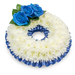 A carefully composed circular wreath photographed from a slightly raised angle, set against a pristine white background that lets every detail read clearly: the central volume is formed by hundreds of pure white chrysanthemums, their soft, plumed petals creating a gently domed surface whose tiny creamy centers add a whisper of warmth. On the upper left, three striking deep blue roses form a concentrated accent-each rose with velvety, closely folded petals in a rich sapphire tone, nestling into clusters of dark, glossy foliage that deepen the colour contrast and draw the eye. The wreath is edged on both the outer and inner circumference with a pleated cobalt satin ribbon; the ribbon's sheen and ruffled texture frame the flowers, giving the design a formal, finished silhouette. A soft shadow under the wreath conveys depth and the impression of careful studio lighting, appropriate for a tribute intended for a funeral or memorial service. As a florist serving families in Gerrards Cross and neighbouring Chalfont areas, and through Flower Delivery Gerrards Cross, I often prepare such pieces for quiet services near the railway station or on the village High Street; the palette speaks of dignity and comfort. There is a subtle sensory balance here-chrysanthemums' delicate scent and cushion-like texture paired with the roses' velvety richness and the cool gloss of the ribbon-resulting in an arrangement that feels at once traditional, personal, and consoling to those saying goodbye.