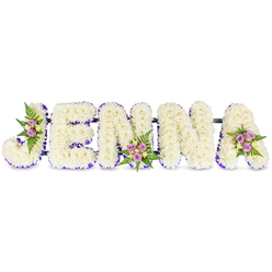 This personalized sympathy piece from Flower Delivery Gerrards Cross presents the name JENNA in bold, three-dimensional block letters, each letter lovingly formed from hundreds of small, snowy-white chrysanthemums or similar compact blooms to create a cloud-like, serene surface. The white flowers are artfully shaded by a thin, deep purple ribbon or trim that follows the inner edges of every letter, giving each form crisp definition while maintaining a gentle colour harmony. In three carefully chosen spots-on the sweeping curve of the J, at the lower centre of the second N, and on the upper right of the A-small clusters of blush-purple spray roses mingle with a few cream or pale yellow roses, their petals carrying a soft, nostalgic scent. Dark green fern fronds cradle these mini bouquets, adding texture and a living depth against the white backdrop. Sleek, unobtrusive dark connecting bars link the letters, ensuring the tribute reads as a unified display suitable for laying beside a casket, standing at a funeral service, or resting at a reception in local halls near Gerrards Cross station. The lighting in the photograph is gentle and diffuse, throwing soft highlights across the petal surfaces and accentuating the delicate craftsmanship. As a memorial arrangement this custom piece balances solemnity with tender warmth, the purple trim offering a touch of individuality and the rose clusters acting as small points of remembrance-an expertly executed farewell available for local delivery across Gerrards Cross, Burnham, and Iver.
