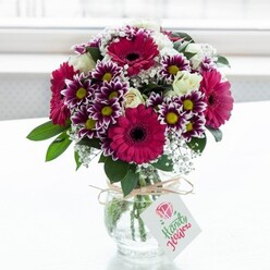 A lush, professionally arranged bouquet is captured in a moment of soft daylight, standing in a squat, clear glass vase with subtle texture that gently distorts the stems within. The flowers form a rounded, abundant dome of colour, where bold fuchsia and magenta gerbera daisies immediately command attention, their dark centres creating a striking focal contrast. Around these, daisy-like chrysanthemums unfurl in intricate layers, each petal shaded from deep purple at the tips to luminous white nearer the centre, where bright yellow eyes catch the light like tiny suns. Nestled between the bolder blooms, creamy white roses appear in varying stages of openness, some just beginning to unfurl, others cupping the light in their layered petals, adding a note of romantic softness. Delicate tufts of white baby's breath, or gypsophila, weave through the arrangement, lending a fine, misty texture that lightens the overall look and accentuates the fullness of the design. Glossy green foliage peeks out from beneath and between the blossoms, grounding the vivid colours with a fresh, natural base. The vase is cinched with a loop of rustic twine tied into a simple bow, from which hangs a small rectangular white tag bearing a stylised pink rose graphic and gentle script, hinting at a handwritten dedication or message. Behind the bouquet, the background fades into a bright, softly blurred white, reminiscent of a sunlit window in a quiet Gerrards Cross home, perhaps overlooking West Common or a leafy side street off Oxford Road. The entire composition radiates cheerful celebration and thoughtful gifting, like the sort of arrangement a local florist would deliver for birthdays, thank you gestures, or new home celebrations around Gerrards Cross and the surrounding Buckinghamshire villages.