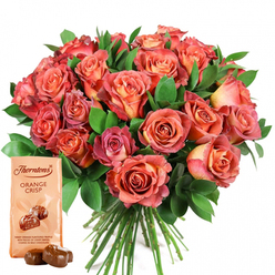 As a Gerrards Cross florist with deep roots in the community, I always strive for an arrangement that honours both form and feeling, and this vibrant gathering truly shines. The densely packed bouquet-brimming with roses of coral, peach, and rich salmon-seems to glow from within, each flower unfurling with soft ombre transitions that echo the kind of sunset you might catch over West Common on a summer evening. The velvety petals are complemented by glossy, dark green foliage, possibly fragrant eucalyptus, their freshness recalling morning dew on a walk through East Common's trails. There's a reassuring softness in the textures: the roses' plush blooms and the leafy greenery invite you to linger. Off to the lower left, Thorntons Orange Crisp chocolates-pouch upright, truffles shining in natural light-promise a sweet finish, their tangy orange notes reminiscent of local summer fêtes. The overall presentation feels sincere and generous, perfect for marking milestones, from a celebrated birthday on Camp Road to anniversaries shared in a Gerrards Cross garden, or just to offer comfort and delight when words aren't quite enough.