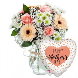 Set against a spotless white backdrop, this Mother's Day composition feels like a moment of quiet celebration captured in still life. A generous bouquet of fresh flowers sits in a clear glass vase, its jar-like shape and simple lines softened by a loose tie of light brown twine around the neck, echoing the relaxed charm of a Gerrards Cross kitchen table on a Sunday morning. The bouquet itself is lush and rounded, filled with pastel blooms that seem to glow softly. Several peach-pink roses, fully open yet still plump with life, display their classic spiral of petals, each petal overlapping the next like folded satin. Around them, peach-orange gerbera daisies with deep, dark centres stretch out their wide, even petals, offering a clean, modern contrast to the romance of the roses. Fine sprigs of white baby's breath weave among the larger blooms, creating a cloud-like haze, while smaller daisy-style chrysanthemums with pale green centres tuck into the gaps, lending texture and a fresh garden feel. Deep green leaves and foliage surround the flowers, framing their colours and suggesting they've been arranged with care by a local florist who knows how to balance shape and shade. Behind the flowers, a small white greeting card peeks out, its colourful writing just visible but not fully readable, like a private message meant for one special mum. At the front right, leaning against the vase, a prominent heart-shaped foil balloon adds a playful, festive note. Its white surface is dotted with scattered metallic gold and silver spots, and in the centre a peach heart carries the message "HAPPY Mother's DAY" in bold black print and flowing calligraphy. Together, the bouquet and balloon create a tender, uplifting gift, perfectly suited to brighten a Mother's Day morning anywhere in Gerrards Cross.