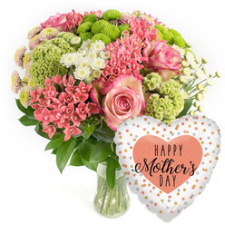 Set against a bright white, uncluttered background, this image focuses on an exuberant Mother's Day display that feels both stylish and heartfelt, as if it has just been arranged by a careful florist in Gerrards Cross. At the centre is a full, domed bouquet presented in a clear glass vase, its stems neatly cut and immersed in water, giving a sense of cool freshness. The composition is anchored by several generous pink roses, their petals layered in soft spirals with gently darker pink edges that appear almost hand-painted. Clusters of coral-pink kalanchoe-like blossoms fill the gaps between the roses, creating a tapestry of tiny, waxy florets that add visual richness. Threaded throughout are vivid lime-green button chrysanthemums and plush, frilly chartreuse accents that bring a contemporary twist, their rounded shapes and tactile surfaces drawing the eye the way new growth might in a Bulstrode Park flowerbed. Small white spray chrysanthemums, reminiscent of daisies, are scattered through the design, introducing a quiet brightness that keeps the palette light and uplifting. Dark, glossy foliage supports the flowers, creating a deep green nest that makes the pinks, corals, and greens glow. To the lower right of the arrangement, a heart-shaped foil balloon leans in like a cheerful companion. Its surface is white with a sprinkling of irregular metallic dots in gold, silver, and soft peach, catching the light in gentle flashes. At its centre, a bold peach-coloured heart frames the message "HAPPY Mother's DAY" in contrasting black script and block letters. The scene suggests a complete Mother's Day surprise, ideal for same-day delivery to a home near East Common or a cosy house off Oxford Road.