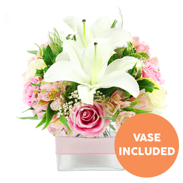 This image shows a compact yet luxurious bouquet that feels perfectly at home on a light-filled table in Gerrards Cross, perhaps overlooking West Common. Dominating the composition are pristine white Oriental lilies, two of them fully open with broad, sculptural petals and prominent dark reddish-brown stamens, and a third lily bud just beginning to unfurl. Nestled closely around these statement blooms are numerous pink roses, ranging from tight, promising buds to generously open flowers whose layered petals form soft, romantic whorls. Between the roses, slender stems of pink and white alstroemeria extend their trumpet-shaped blooms, their speckled yellow throats adding gentle warmth and movement to the design. Fine clusters of white baby's breath drift through the bouquet like mist, filling small gaps and bringing a sense of airiness that keeps the arrangement from feeling heavy. The flowers are supported by a rich base of greenery: broad, glossy leaves catch the light, while more delicate, feathery foliage adds texture and a natural, garden-like feel. Everything is gathered into a low, square glass vase, crystal clear so that the flower stems are visible, giving an honest, freshly arranged appearance. Around the middle of the vase, a pale pink satin ribbon is wrapped smoothly and tied at the back, adding a soft, decorative band of colour that echoes the roses. In the lower right corner, an orange circular graphic with bold white capitals clearly states VASE INCLUDED, making it evident that this is a complete, ready-to-display gift. The bouquet radiates tranquillity and gentle joy, ideal for birthdays, get-well wishes, or simple thinking of you deliveries across Gerrards Cross and the surrounding lanes.