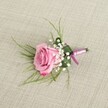A single blush-pink rose boutonniere rests on a softly textured, light beige linen cloth, captured from a gently elevated angle that lets you see every petal and ribbon detail. The rose is semi-open rather than fully blown, its velvety petals curling delicately at the edges, with colour that drifts from a pale, romantic pastel in the centre to a richer, more vivid pink along the outer rim. Tiny, cloud-like clusters of creamy white baby's breath nestle closely around the base of the bloom, their starry flowers adding a lace-like softness and a classic wedding feel. Behind the rose, fine emerald-green blades of grass-like foliage arc outward in slim, wispy lines, creating a light, airy frame, while a single broader, darker green leaf is folded neatly to cradle the stems, giving the arrangement a sense of structure and polish. The stems are tightly bound with a lustrous satin ribbon in tones of lavender-pink and deeper magenta-purple, tied in a modest knot that adds just enough shine without overwhelming the rose. The natural, woven grain of the linen beneath is visible and gently illuminated by soft, even light, offering a calm, neutral backdrop that makes the boutonniere's colours seem especially fresh and romantic. It looks like the sort of lapel flower a groom or prom guest might wear to a celebration in Gerrards Cross, perhaps walking from the station towards a reception near East Common, its understated elegance perfectly in tune with a refined, modern countryside gathering.
