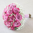 Set diagonally across a softly creased, off-white fabric, this bouquet feels like a moment of romance captured in stillness. The focal point is a cluster of plump roses, around a dozen in number, each bloom displaying a sophisticated gradient of colour: inner petals in muted lavender-pink gently deepening to vivid fuchsia and intense magenta at the tips. The roses are not fully blown, holding a graceful semi-open form that reveals layers of tightly swirled petals, suggesting both freshness and longevity. Encircling this vivid heart is a generous ring of white baby's breath, its myriad tiny blossoms creating a light, airy veil that softens the stronger hues and adds texture, like morning mist over the commons in Gerrards Cross. Discreet flashes of dark green leaves anchor the palette and lend a natural balance, keeping the composition from feeling overly sweet. The stems are gathered precisely and extend towards the lower right corner, wrapped in pale purple satin ribbon that feels smooth and cool to the touch. Over the ribbon, a spiral of delicate white pearls catches small points of light, echoing bridal jewellery and giving the bouquet an unmistakably ceremonial character. The overall mood is calm, intimate, and celebratory, as though this arrangement has just been set down before a wedding or engagement photo. Crafted with the care you'd expect from an experienced Gerrards Cross florist, this design is ideal for illustrating local weddings, anniversaries, or elegant same-day flower delivery in and around the village.