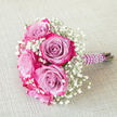 A romantic, small bouquet lies diagonally across a pale, woven beige fabric, the gentle texture beneath it echoing the softness of the blooms. Several bi-colour roses form the focal point, each one with a cool lavender or light lilac heart that gradually deepens into rich magenta-pink along the outer petal edges. The petals are densely layered, creating a full, rounded shape that looks plush to the touch, like a carefully crafted rosette. Between the roses, clouds of white baby's breath spill outward, their multitude of tiny blossoms resembling soft, scattered stars against the richer hues of the roses. This delicate gypsophila adds both fullness and airiness, balancing the boldness of the rose colours with a sense of innocence and purity. The stems are neatly bound in a deep purple satin ribbon, wrapped so smoothly that the fabric appears almost liquid, and then adorned with a spiral of glossy white faux pearls that trace down the handle. The pearls add a classic, almost vintage refinement, making the bouquet feel especially suited to a bridesmaid or young bride celebrating in Gerrards Cross, perhaps on a quiet lane near the Memorial Centre or close to the station. The lighting is calm and even, with no harsh shadows, allowing the eye to linger on the colour gradients and the contrast between the rich florals and the simple backdrop. It feels like a bouquet moments before being carried at a local ceremony, ready for wedding photos or same-day delivery for an intimate celebration.