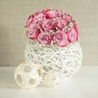 A romantic still life shows a lush, perfectly rounded bouquet of soft pink roses and white baby's breath, resting in a contemporary woven white vase on a clean white surface. Around eighteen to twenty roses form a tight dome, each bloom opening generously so that the petals reveal a gentle ombré of colour, from a pale blush at the heart to a deeper, almost fuchsia pink along the outer edges. Clouds of tiny white gypsophila flowers are threaded between the roses, light and feathery, giving the bouquet a floating, cloud-like softness that feels both modern and timeless. The flowers sit in a spherical vase crafted from criss-crossing white strands, reminiscent of painted wicker or rattan, with open gaps that let the soft light filter through and cast subtle shadows. In front of the vase, two natural decorative spheres in light beige tones echo the woven texture; their thin wooden strips loop and weave into airy globes, the larger sphere set slightly behind the smaller for a gentle sense of depth. The entire arrangement is placed against a warm beige or cream backdrop, similar to the soft tones you might see in a calm Gerrards Cross sitting room overlooking West Common, giving the scene a peaceful, homely charm. The light in the image is bright yet diffuse, as if from a nearby window on a mild spring morning, illuminating every petal edge and strand of wicker. The composition feels like a carefully styled table or mantelpiece display, ideal for a quiet anniversary at home or a thoughtful surprise delivered to a loved one in Gerrards Cross, capturing that mix of freshness, elegance and understated celebration that so many local clients seek for intimate occasions.