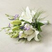 Lying gently across a subtly textured off-white linen, this sophisticated bridal bouquet is a testament to the artistry and heart of Gerrards Cross florists. The arrangement is dominated by stately white Oriental lilies, their open trumpet blooms and elegant petals touched by fine berry-hued freckles and golden-hued stamens. These stunning focal flowers are joined by rippling lisianthus blossoms, their snowy petals edged dramatically in violet, weaving a lively accent through the bouquet. Tight buds of lisianthus nestle among the fullness, suggesting a promise of blooms still to unfold, much like a spring morning in the green spaces near Bulstrode Park. Accents of slender purple veronica add vertical lines and deeper purple depth, while airy gypsophila sprigs lend a gentle, cloudlike softness that softens the entire composition. The bouquet is framed by an abundance of lush greenery: broad leaves supporting the lilies, and trailing sprays of feathery asparagus fern reminiscent of the lush borders in local gardens. Neatly gathered stems, finished with a white ribbon, mirror the elegant simplicity of a Gerrards Cross wedding. Under a soft wash of even light, each bloom and leaf is revealed in delicate detail-an arrangement that embodies not just natural beauty but hopefulness and new beginnings. The bouquet, ideal for bridal parties, anniversaries, or special celebrations, is available for thoughtful same-day delivery throughout the Gerrards Cross community, carrying elegance and joy wherever it travels.