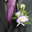 A close-up photograph shows the upper torso of a man dressed for a wedding in Gerrards Cross, his formal attire transformed by a carefully crafted buttonhole that becomes the quiet star of the image. He wears a dark charcoal grey suit jacket, sharply tailored with a clean lapel that catches a soft, natural light, perhaps like the gentle brightness you get on a calm morning walking past the green of East Common. Underneath sits a light grey checkered dress shirt, its subtle pattern adding texture without distraction. At the centre of the scene is a lustrous magenta silk tie, smoothly knotted, whose vivid colour sings against the darker fabric and leads the eye straight to the boutonniere pinned neatly on the left lapel. The floral design features a white lisianthus bloom edged with deep, ruffled purple, its petals layered like delicate watercolor strokes, and a second bloom or half-open flower echoing the same tones. Clustered around are small, closed white buds hinting at new beginnings, a slender vertical spike of dark indigo-purple veronica adding height, and tiny whispers of white filler flowers, similar to baby's breath, that soften the overall look. Lush green foliage frames the flowers: feathery sprigs, smooth emerald leaves, and fine loops of decorative green grass that lend a modern, slightly architectural twist. At the base, a small white satin ribbon is tied into a flawless bow, neatly wrapping the stems and giving the arrangement a refined, timeless finish. The whole image radiates quiet joy and poised anticipation, the sort of thoughtful detail you might choose from a trusted local florist when planning a ceremony at a favourite church or venue in Gerrards Cross.