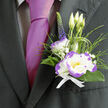 This boutonnière feels at once timeless and subtly contemporary, a small composition that carries a whole day's worth of intention. At its centre sits a white lisianthus, the petals delicate and silk-like, each edged with a striking violet-purple that creates a crisp, dramatic outline when caught by late-afternoon light. Nestled close is a white dianthus with lively purple ruffles, its textured petals harmonising with the lisianthus while offering a softer, more playful counterpoint. Three slender lisianthus buds reach up behind the main bloom, the promise of unopened petals mirroring the optimism of a celebration, and a single deep purple veronica spike gives the design height and a flash of indigo.

Feathery grassy loops and glossy dark-green foliage cradle the blooms, their tactile movement lending a casual grace, while tiny clusters of gypsophila breathe air into the arrangement - like scattered stars against a soft sky. The whole is bound with a crisp white ribbon, neat and reassuring, and set against the lapel of a dark suit so the magenta tie beside it becomes part of the visual story: refined, warm, and slightly mischievous. The bouquet carries a light garden scent that feels like a calm companion through vows, speeches, and photographs.

As a local florist I often prepare pieces such as this for ceremonies around Gerrards Cross station and nearby Beaconsfield, and Flower Delivery Gerrards Cross can arrange same-day hand delivery across the town when a thoughtful finishing touch is needed. It's the kind of boutonnière that feels personal, steadying, and carefully made.