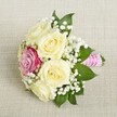 A petite, intricate bouquet rests gracefully on a textured cream fabric, emblematic of Gerrards Cross's intimate celebrations. Five creamy roses, their petals soft and slightly open, create a gentle, enveloping heart to the composition. Off-centre, a vivid magenta-pink rose stands out, a joyful burst of color that brings energy and distinction to the otherwise subtle bouquet. Wisps of white baby's breath fill the spaces between, their delicate blossoms giving the arrangement a cloud-like, romantic quality. Deep, vibrant green leaves peek from beneath the blooms, grounding the colors and offering a fresh accent that mirrors the verdant spaces of Gerrards Cross, such as the Commons or East Common. The bouquet's stems are securely bound with textured pink satin ribbon and entwined with a strand of luminous pearls, the finishing touches adding sophistication and a hint of sparkle reminiscent of a candlelit evening wedding. Bathed in natural daylight, the arrangement showcases every nuance of the petals and foliage, as if inviting the viewer to touch and inhale their subtle floral fragrance. Crafted with love and precision, this bouquet encapsulates the spirit of celebration, making it the perfect floral accent for weddings, anniversaries, or heartfelt gatherings throughout Gerrards Cross.