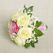 A delicately styled small bouquet lies on a soft, light beige textured background, reminiscent of finely woven linen you might find on a beautifully laid table in a Gerrards Cross home. The arrangement is intimate yet refined, built around a cluster of five creamy white or ivory roses, each one fully open, with layered petals curving outward in gentle, rounded waves. Nestled near the front-left of the design, a single vivid fuchsia-pink rose introduces a bold accent of colour, its saturated hue standing out against the understated whites, like a vibrant flower bed brightening a quiet street off Oxford Road. Scattered generously between the roses are sprays of baby's breath, their myriad tiny white blooms forming delicate, cloud-like clusters that add both softness and lightness to the bouquet. Surrounding the flowers, deep green pointed leaves-likely ruscus or salal-frame the composition, their clean lines and glossy surfaces providing structure and contrast, ensuring the roses remain the clear focal point. The stems are neatly aligned and enclosed in a bright pink satin ribbon that wraps snugly to form a graceful handle. Over this, small iridescent white pearls are arranged in tidy horizontal bands, the beads catching the light and adding a sense of luxury and celebration. Photographed from a slightly raised, close-up angle in even, flattering light, the bouquet's textures and colours appear crisp and true. It feels perfectly suited for use as a corsage, boutonniere, or mini centerpiece for a wedding reception in one of Gerrards Cross's local venues, or as a special detail for a prom or engagement dinner, carrying with it a mood of romance and thoughtful elegance.