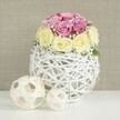 A delicately styled floral centerpiece is showcased against a softly textured beige backdrop, the sort of gentle, neutral wall you might find in a tastefully renovated Gerrards Cross home. At the centre is a beautifully rounded bouquet of around a dozen roses, each bloom chosen for its freshness and subtle colour. The upper portion of the arrangement is composed primarily of pastel pink roses that verge on lavender, their petals layered and slightly ruffled, some still tightly furled while others unfurl into soft spirals. Encircling the base of this pink crown is a neat ring of creamy yellow and ivory roses, forming a subtle band of warm light that stabilises the design and draws the eye around the dome. Between the roses, fine stems of white baby's breath weave in and out, their tiny, star-like blossoms creating a delicate lace that adds lightness and volume. The flowers are set into an unusual white spherical vase made from thread-thin wicker or branches, painted white and shaped into an open, airy globe that resembles a decorative lantern. Its intricate intersections give a sense of craftsmanship and texture. On the white surface beside it rest two smaller decorative spheres made from pale natural rattan, their open-weave design harmonising beautifully with the main vase while adding a relaxed, organic note. Soft, even lighting illuminates the arrangement, enhancing the gentle tones and clean lines without harsh shadows. The entire composition suggests a carefully curated wedding table, engagement celebration, or special family gathering in Gerrards Cross, where understated elegance and handcrafted details help create an atmosphere of shared joy and cherished memories.