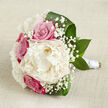 Displayed against a pale, slightly textured beige backdrop that recalls natural linen, this hand-tied wedding bouquet feels both luxurious and effortlessly relaxed, like something carried to a ceremony in Gerrards Cross on a soft spring morning. At the centre is a single large creamy white peony, fully open and plush, its many ruffled petals forming a round, cloud-like focal point. Encircling it are several fuchsia-pink and dusky blush roses, their petals tightly layered in perfect spirals, giving the arrangement a touch of classic romance and depth of colour. Threaded throughout are clusters of fine white baby's breath, creating a delicate lace of tiny blooms that add airiness and movement. Deep, polished green leaves, broad and slightly curled, frame the flowers from beneath, guiding the eye along the bouquet's gentle curves and grounding the pastel tones with rich greenery. The stems are carefully aligned and bound with a sleek white satin ribbon, wrapped firmly but elegantly to form a smooth handle that suggests the bouquet is meant to be held throughout a wedding day. Soft, even lighting caresses the petals, revealing their gentle sheen and natural veining without any harsh contrast, so the entire design appears calm, fresh, and thoughtfully composed. The bouquet feels perfectly suited to a ceremony near East Common or a reception close to Gerrards Cross station, bringing a sense of romance, refinement, and local floral craftsmanship to the occasion.