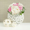 A sophisticated floral centerpiece glows against a soft beige backdrop, featuring lush white peonies and vivid fuchsia-pink roses arranged in a compact, perfectly rounded design that recalls an elegant bridal bouquet in Gerrards Cross. The peonies sit prominently at the heart of the arrangement, their generous, ruffled petals forming creamy domes that seem almost weightless, while the roses encircle them in saturated pink, each petal curling outward with a sense of quiet drama and romance. Threaded throughout the blooms are fine sprays of baby's breath, their tiny, cloud-like flowers catching the light and adding a dreamy, lace-like veil around the main blossoms. At the base, smooth, deep green leaves arc gently around the bouquet, framing it with a polished, natural border and hinting at freshly cut stems prepared by a skilled local florist. The flowers are presented in a distinctive white vase made from delicately interwoven strands that create an open, basket-like lattice, giving the impression of a modern sculpture more than a traditional container. You can see glimpses of the stems and shadows through the gaps, adding an intriguing sense of dimension and movement. On the clean white surface below, two decorative woven balls in pale, straw shades rest nearby-simple yet refined accents that echo the vase's texture and suggest thoughtful styling for a wedding table in a Gerrards Cross venue or a stylish home just off Oxford Road. The overall mood is one of refined celebration, ideal for marking engagements, anniversaries, or intimate receptions across the local area.