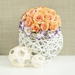 A sophisticated floral centerpiece is arranged on a glossy white surface, evoking the feel of an intimate reception table at a Gerrards Cross celebration. At its core sits a perfectly shaped dome of roses in muted shades of peach, apricot, and soft salmon, the blooms packed so closely together that their velvety petals create an almost seamless, cushion-like sphere. Tucked gently around the base is a ring of white lisianthus, each blossom edged with a band of rich purple and violet, giving the impression of hand-trimmed petals and adding a refined contrast to the warm rose tones. Threaded between these are fine sprays of white baby's breath, their tiny flowers creating a light, airy mist that softens the edges of the arrangement. The flowers are held in a broad, bulbous container crafted from white woven wicker or rattan, its open latticework structure forming a sculptural vessel through which light and shadow interplay. Resting in front of the main arrangement on the same white surface are two decorative spheres, one slightly larger than the other, made from slender, natural beige strips of wood woven into loose, airy orbs that echo the basket's texture. Behind the display, a gently textured beige fabric backdrop, reminiscent of linen or burlap, offers a warm and neutral setting that lets the subtle colours and intricate details shine. The overall mood is one of calm sophistication and quiet romance, making this design ideal for wedding tables, anniversary dinners, or elegant home styling around Gerrards Cross, with the kind of artisan care a local florist brings to each arrangement.