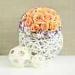 In a soft, inviting composition that feels both modern and handmade, a compact dome of peach to apricot roses forms the visual heart of this arrangement - roughly two to three dozen blooms gathered so their textures read like layers of silk and suede. Several roses are fully open, their concentric petals catching a gentle light, while others remain in tight, hopeful buds; the balance creates a sense of freshness and the slow unfolding of fragrance. Encircling the lower circumference of the rose mound are white Lisianthus blossoms, their petals quietly creamy yet edged in vivid purple, a painterly contrast that adds a touch of refined drama. Delicate sprigs of white Gypsophila are woven through the composition, lending a lacy, breathy halo that brightens the apricot tones. The flowers are presented in a rounded white woven vessel - an airy lattice of reeds or synthetic wicker - its open, sculptural form bringing a contemporary, artisanal sensibility. On the left of the arrangement sit two woven wooden or bamboo spheres, one taller than the other; their pale, tactile curves echo the basket's pattern and introduce a natural, Scandinavian-inspired accent. The backdrop is a neutral, linen-coloured wall with soft texture and shallow depth of field, keeping attention on the floral forms. The lighting is calm and even, suggesting a quiet morning on Gerrards Cross near the station or a polished window-side in a High Street home. This bouquet reads as thoughtful and celebratory, suited to anniversary tables, bridal showers, or as a carefully chosen gift for someone who loves understated, modern elegance.