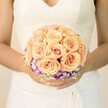 A close-up, chest-level photograph shows a bride standing quietly, her torso framed against a soft, creamy background that feels like gentle daylight spilling into a historic Gerrards Cross venue. She wears a classic white or ivory wedding gown with delicate lace tracing the bodice and a modest V-neckline, the fine pattern almost like frosted leaves. Soft, flowing folds of fabric fall from her waist, adding a sense of movement and tradition. Cradled in her hands is a perfectly round bridal bouquet, clearly the focal point of the image and crafted with great care by an experienced florist. The bouquet is densely packed with luxurious roses in warm shades of peach, apricot, and blush pink, every petal neatly cupped and layered, giving the impression of sumptuous, velvety spirals. Around the lower edge of this rose dome, a bold ring of deep purple blooms with ruffled petals tipped in white-likely lisianthus-forms a dramatic contrast, as if a soft halo of twilight encircles the warmer centre. Tiny clusters of white filler flowers, reminiscent of baby's breath, are tucked between the purples and peaches, lending a cloud-like lightness and a hint of countryside charm, as though the stems had just been gathered near West Common. The bride's manicure is understated and elegant, with pale, neutral nails and one thumb catching the light with a subtle shimmer, mirroring the quiet sparkle of the day. The overall impression is of romance, composure, and anticipation-a timeless bridal bouquet, perfect for a wedding in Gerrards Cross, whether in a leafy garden setting or a classic church ceremony, captured at the moment just before she walks down the aisle.