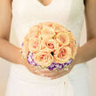This arrangement presents itself as a study in gentle harmony-apricot-peach roses clustered at the heart, each blossom dense and carefully shaped so the bouquet reads as a single, luminous form. Around that warm centre, a ring of purple-tipped and white lisianthus creates a subtle, cool border that gives depth and a hint of romantic contrast. Fine, lacy filler blooms weave through the composition, their airy texture softening the overall silhouette and catching the light in tiny highlights. The petals show delicate veins and a cream-to-peach gradient that the photographer has captured in soft, diffuse daylight, producing a tender translucence at the edges. The tactile experience is immediate in description: the velvet smoothness of rose petals, the papery crispness of lisianthus frills, and the faint yet uplifting scent of fresh roses with green notes from foliage. As a local florist who prepares bouquets for ceremonies and celebrations around Gerrards Cross station, the Chalfonts and nearby Denham, I balance visual impact with comfortable weight and secure binding so the bouquet is both beautiful and easy to hold. Flower Delivery Gerrards Cross carefully positions each bloom to read well in photographs taken on the High Street or in village gardens, while keeping the palette intentionally soft for a timeless bridal look. This bouquet fits a bride seeking restrained luxury-poised, tactile, and quietly radiant-designed to be both held in the moment and remembered with warmth.