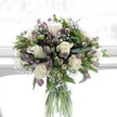 A rounded bouquet of creamy white roses, lavender alstroemeria, and airy baby's breath sits gracefully in a clear cylindrical glass vase, bathed in soft natural light that feels like it's streaming through a Gerrards Cross bay window. The white roses are fully open, their velvety petals unfurling in generous spirals that create a plush, romantic focal point. Around them, clusters of dusky lavender and muted purple alstroemeria buds and half-open blooms introduce gentle colour, their slender petals brushed with deeper purple veining and still hugged in places by green sepals. Wisps of white gypsophila weave between the larger flowers like tiny clouds, softening every edge and giving the arrangement a delicate, mist-like halo. Lush green foliage frames the blooms, from broad, dark leaves that add depth and structure to slimmer stems that arch lightly, guiding the eye around the dome of flowers. Through the transparent glass vase, the neatly trimmed green stems are clearly visible, aligned in fresh water that catches the light and underscores the bouquet's freshness. The whole design feels calm and uplifting, as if it could be resting on a kitchen table overlooking Austenwood Common or a sunlit sitting room just off Packhorse Road. This is the kind of bouquet often chosen to bring quiet comfort after a loss, to offer a gentle congratulations, or to brighten a neighbour's day in Gerrards Cross, its serene whites and lilacs creating a sense of purity, reassurance, and thoughtful care crafted by an experienced local florist.