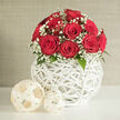 A lush dome of intense crimson roses forms the heart of this arrangement, each flower nestled closely against the next so that the bouquet appears almost like a single, luxurious red cushion. The petals are full and velvety, gently curling back to reveal intricate spirals that speak of freshness and care. Encircling the roses is a delicate fringe of white baby's breath, thousands of tiny blossoms creating a lace-like border that softens the boldness of the red and adds airiness to the design. Just at the base, discreet hints of dark green foliage peek through, giving a natural finish without disturbing the rounded silhouette. The bouquet rises from a striking white woven vase shaped like a globe, its slender bands of wicker or rattan crossing in an open lattice that looks both contemporary and handcrafted. Light passes through the weave and falls onto the white tabletop below, making subtle patterns that enhance the tactile quality of the scene. Two pale, cream-toned wicker balls are arranged casually beside the vase, one slightly larger, both echoing the same interlaced texture and adding a decorative, almost sculptural touch. Behind the display, a smooth wall in muted beige or light grey keeps the focus firmly on the interplay between red, white, and soft natural fibres. The overall mood is serene and romantic, like a carefully set corner in a Gerrards Cross home ready for an intimate wedding gathering or anniversary dinner. It feels like a design a local florist would create for same-day delivery around Bulstrode Way or East Common when someone wants to express deep affection with understated elegance.