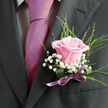 Pinned precisely on a charcoal-grey lapel, this pink rose boutonniere looks like a small painting brought to life. The rose itself is a gentle shade of warm blush, each velvety petal curving outward in smooth, even layers that suggest both tenderness and quiet confidence. Around it, airy sprays of white gypsophila form a delicate halo, their tiny, rounded blossoms hovering like soft snowflakes against the structured fabric of the suit. Slender fronds of greenery stretch upward and outward, adding a graceful line and a hint of woodland texture, their fine needles creating a touchable contrast to the rose's satin-smooth surface. The stems are bound tightly with a lilac satin ribbon, the bow lying flat and refined, offering a polished finish that reflects the formal mood of a wedding or black-tie event in Gerrards Cross. It is easy to imagine the subtle fragrance rising as the wearer walks through West Common or towards a ceremony at a nearby venue: the fresh, green notes of the foliage blending with the romantic scent of the rose. The lighting is soft, likely natural daylight filtering through a window, allowing each shade of pink, white, and green to sit harmoniously together. Crafted with the steady hands of a local florist, this boutonniere becomes more than an accessory; it is a personal emblem of joy, commitment, and celebration, worn close to the heart on a meaningful day in Gerrards Cross.